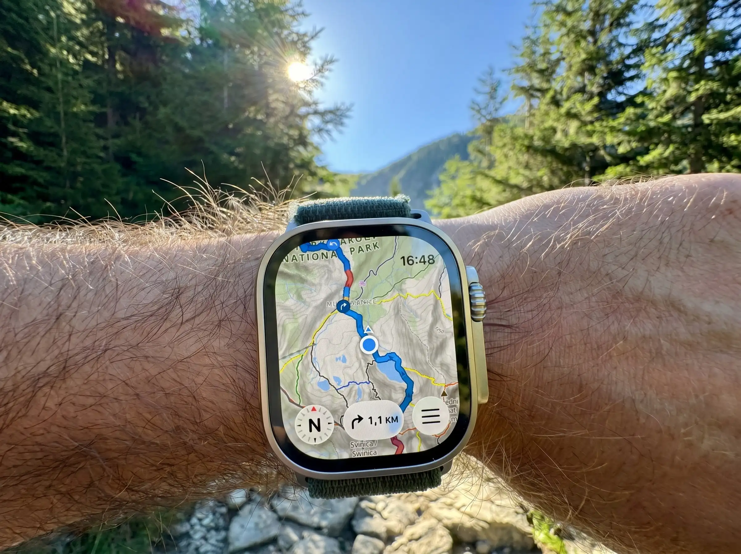 Apple Watch Ultra worn on a wrist in a sunlit mountain forest, showing a WristTopo topographic map with a blue route and the distance to the next turn